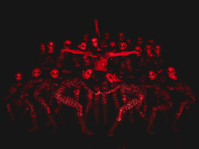 Een groep mensen in donkere kleding poseert dramatisch in een rode, mysterieuze setting.