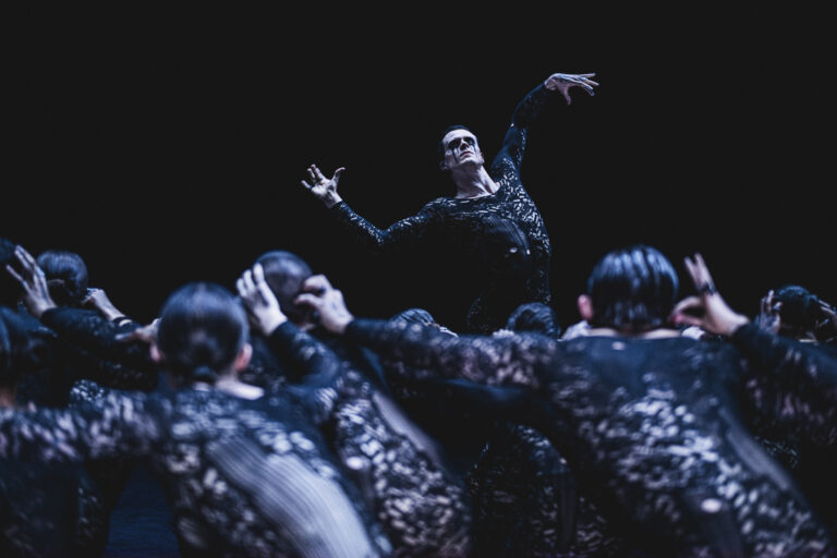 Een danser in zwart kleding voert een expressieve beweging uit, omringd door andere dansers in dezelfde outfits.
