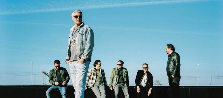 Vier mannen staan buiten tegen een heldere blauwe lucht, gekleed in casual jassen en zonnebrillen.