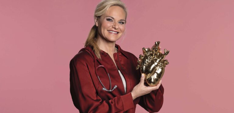 Glimlachende vrouw in rode dokterskleding houdt een grote gouden hartvormige sculptuur vast tegen een roze achtergrond.