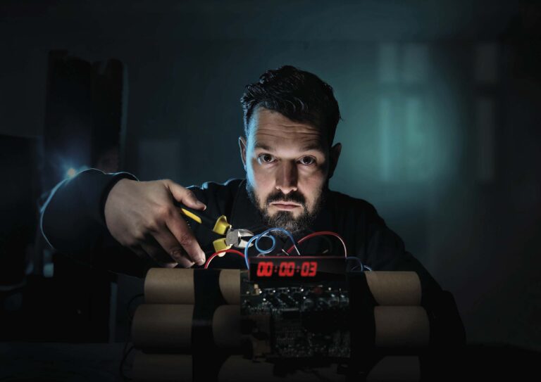 Een man met baard kijkt recht in de camera terwijl hij voor een computer zit in een donker verlichte kamer met blauwgroene verlichting.