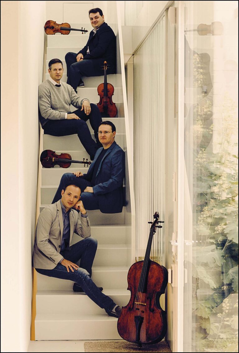 Vier mannen zitten op een trap in een huis, met een gitaar die tegen de leuning rust.