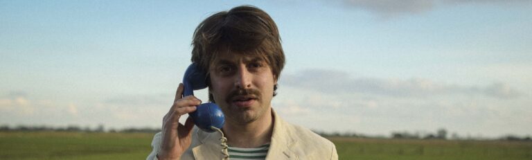 Een man met een snor houdt buiten een blauw bedraad telefoonhoorn aan zijn oor.