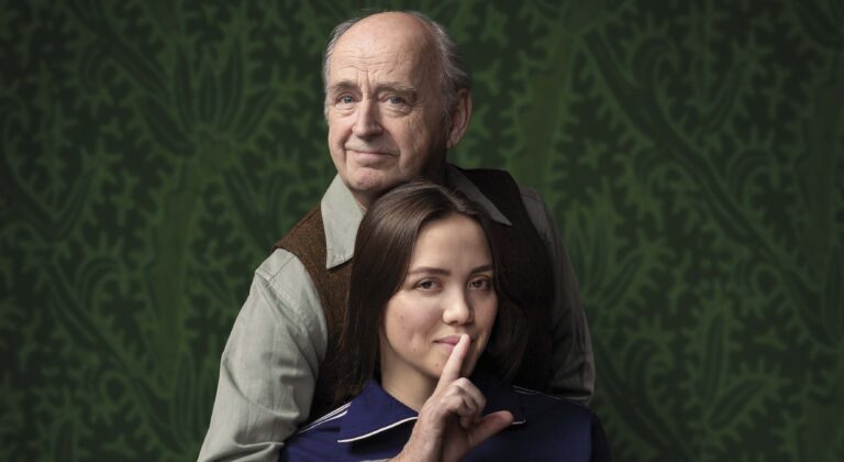 Een oudere man staat achter een jonge vrouw; de vrouw legt haar vinger tegen haar lippen als teken van stilte.