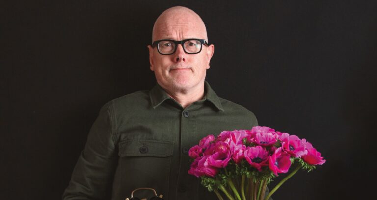 Een persoon met een bril houdt een boeket roze bloemen vast voor een donkere achtergrond.