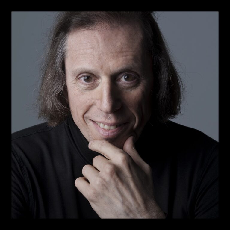 Een man met lang, donker haar glimlacht en steunt op zijn hand, terwijl hij in de camera kijkt.