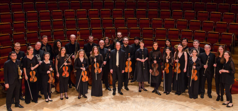 Een groep muzikanten in zwarte kleding staat op een podium met lege rode stoelen op de achtergrond.