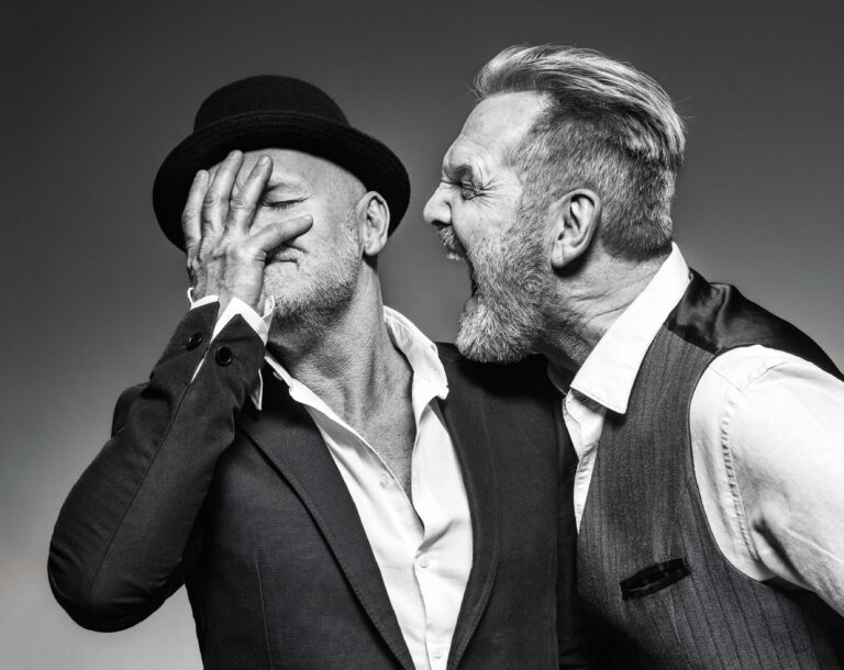 Zwart-wit foto van twee mannen in pak; één met een hoed die zijn gezicht met zijn hand bedekt, terwijl de ander naast hem fluistert.