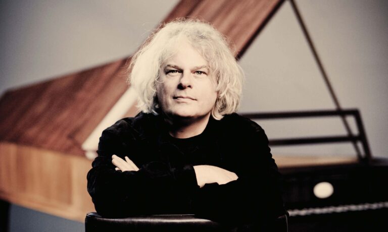 Portret van een oudere persoon met licht blond haar, zwart gekleed en armen over elkaar, voor een geometrische achtergrond.