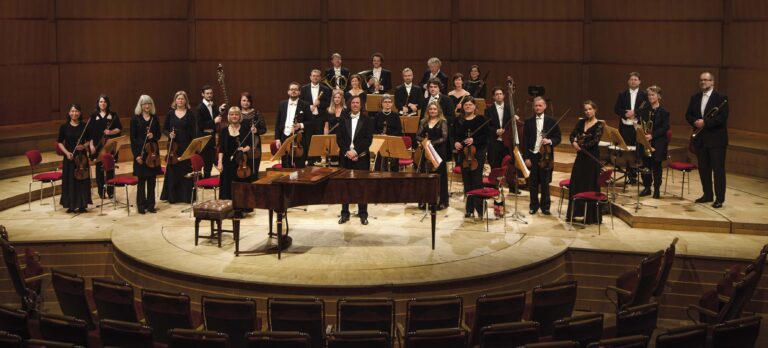 Een symfonieorkest staat op een concertzaalpodium met een vleugel in het midden.
