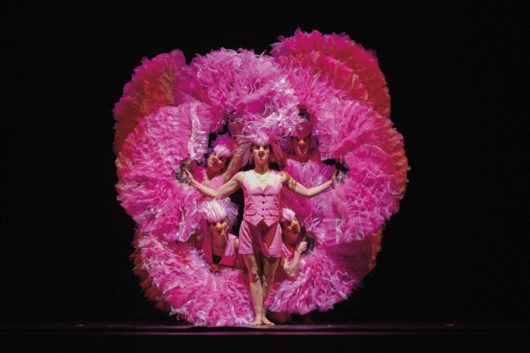 Twee cabaretdansers op een donker podium, met grote roze veerfans die een cirkel achter hen vormen.