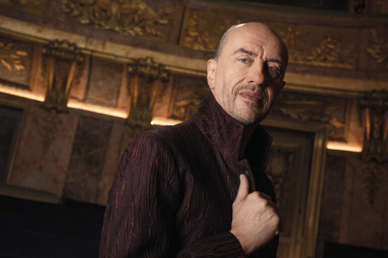 Een man met een korte baard staat in een rijk versierde zaal en kijkt naar de camera.