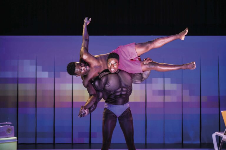 Twee dansers voeren een acrobatische beweging uit op een podium met een kleurrijke achtergrond.