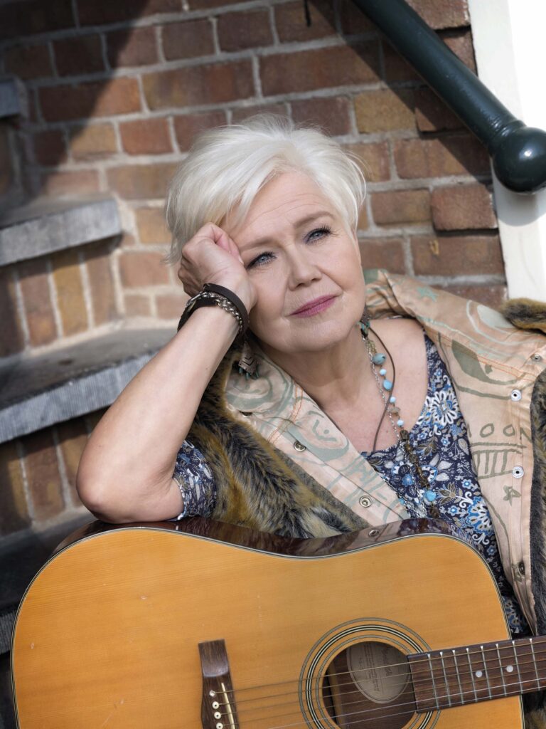 Een vrouw met grijs haar leunt tegen een muur met bakstenen en houdt een gitaar vast, terwijl ze in gedachten verzonken lijkt.