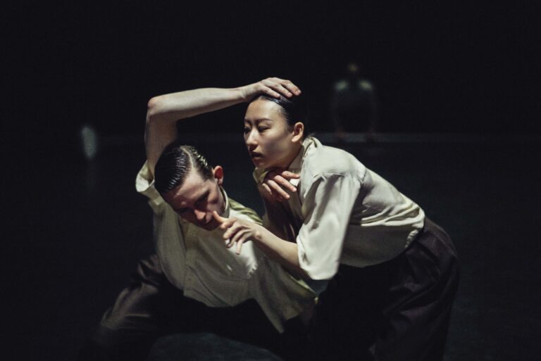 Twee dansers in interactie, met dramatische houdingen in een donkere omgeving.