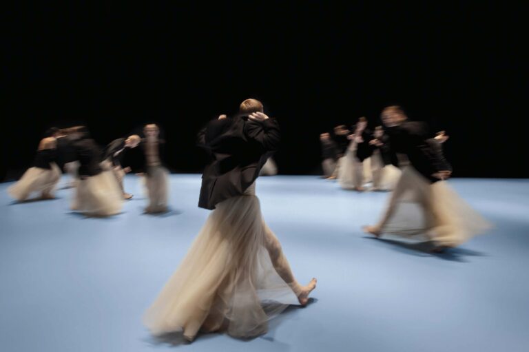 Een groep dansers in elegante kleding beweegt dynamisch over een blauwe achtergrond.