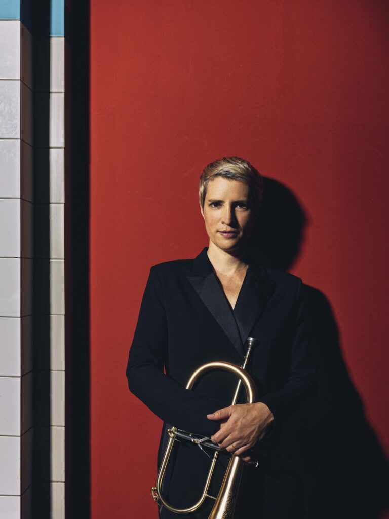 Portret van een persoon met kort, licht haar, gekleed in een zwart jasje, staand tegen een rode muur.