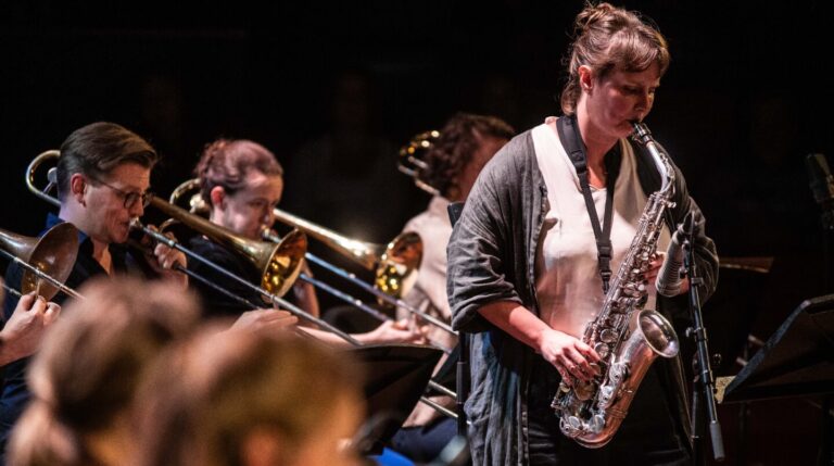 Een muzikante speelt saxofoon terwijl andere leden van het ensemble trombone spelen op de achtergrond.