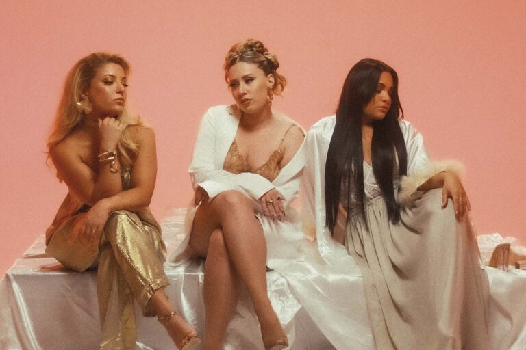 Drie vrouwen poseren op een bed met een pastelroze achtergrond, gekleed in elegante, luchtige outfits.