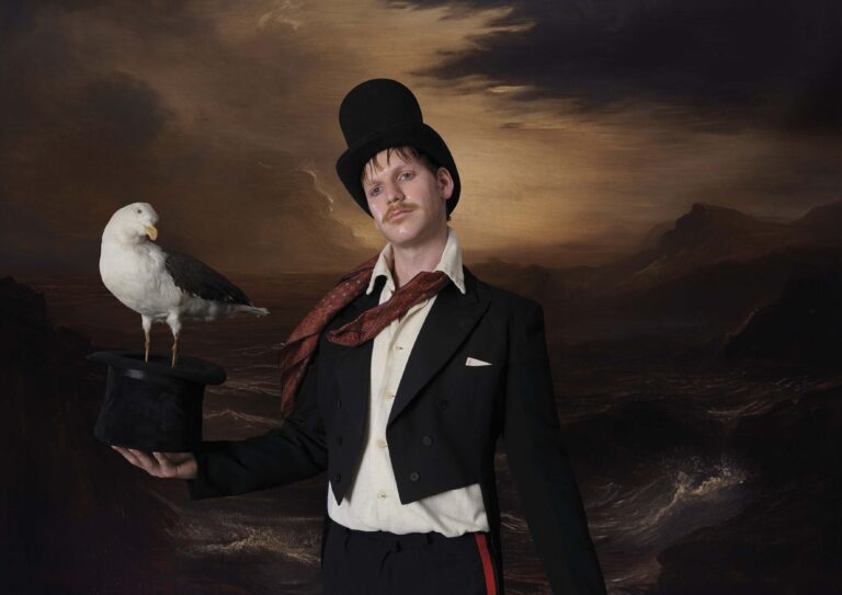Een persoon in een zwart en wit kostuum staat met een hoed en houdt een zeevogel op een hoge hoed vast, met een dramatische achtergrond.