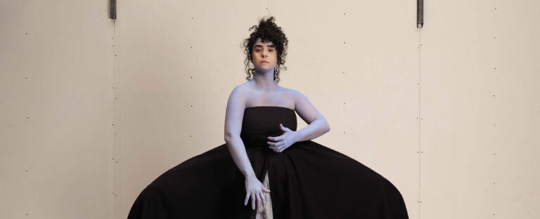 Een persoon met een blauw geverfde huid staat in een zwart, strapless jurk voor een neutrale achtergrond.