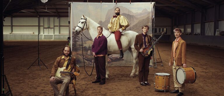 Een groep vier mannen, waarvan er één op een wit paard zit, poseert voor een geschilderde achtergrond in een stal.