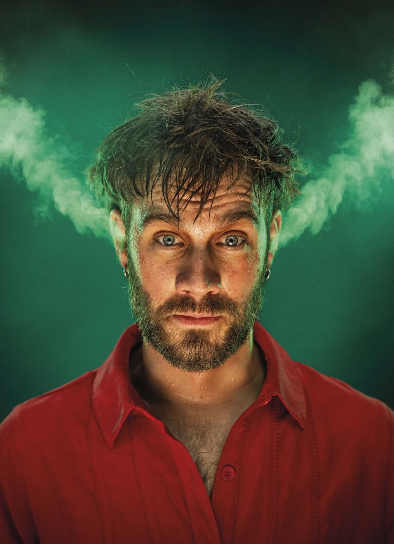 Een man met wilde haren en een baard kijkt recht in de camera, terwijl er dampige rookwolken van zijn hoofd opstijgen tegen een groene achtergrond.