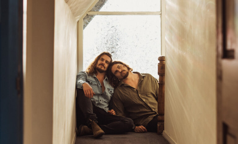 Twee mannen zitten samen op de grond in een gang, met hun hoofden tegen elkaar geleund en een raam op de achtergrond waar water naar beneden stroomt.