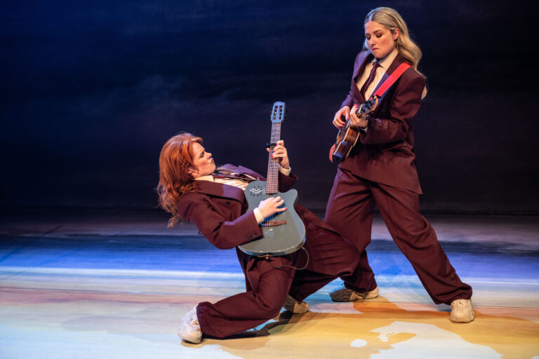 Twee muzikanten in herkenbare outfits spelen gitaar op een podium met een dramatische achtergrond.