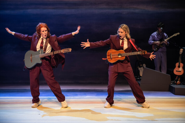 Twee vrouwelijke performers in bordeauxkleurige pakken spelen gitaren op een podium, terwijl een muzikant in de achtergrond meedoet.