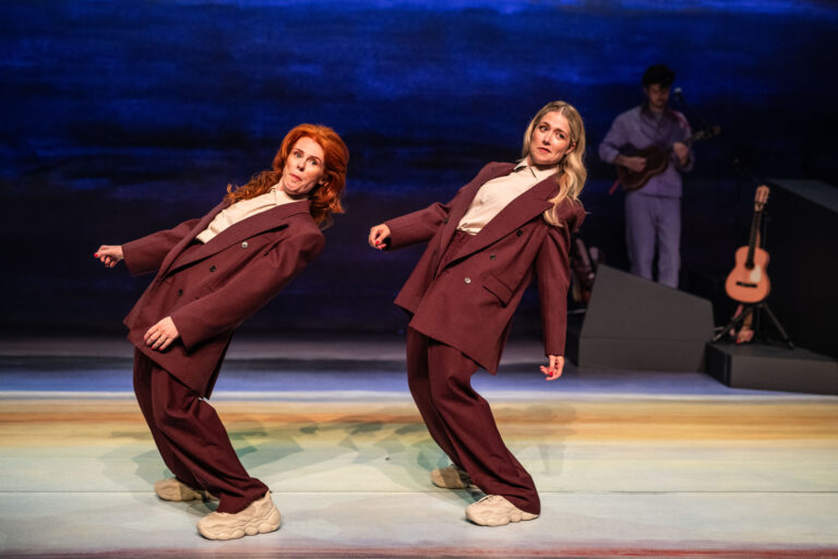 Twee dansers in oversized kleding bewegen synchroon op een kleurrijk podium, met een muzikant op de achtergrond.
