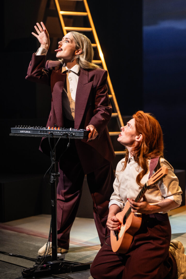 Twee acteurs op het podium, met de ene die een synthesizer bespeelt en de andere die een gitaar vasthoudt, terwijl ze elkaar aankijken.