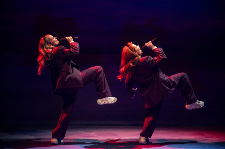Twee dansers in zwarte outfits en witte sneakers voeren een krachtige choreografie uit op het podium, met een kleurrijke achtergrond.