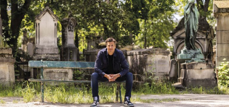 Een man zit op een bank in een park met bomen op de achtergrond.