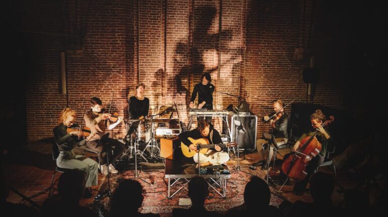 Een muzikale groep in een sfeervolle ruimte met stenen muren, die verschillende instrumenten bespelen terwijl ze zich op een groot tapijt bevinden.