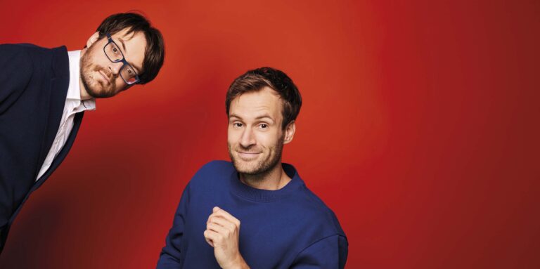 Een man in een blauwe trui glimlacht en poseert speels voor een felrode achtergrond.