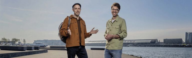 Twee mannen staan buiten bij een wateroppervlak, met verschillende kledingstijlen, en kijken naar de camera terwijl ze glimlachen.