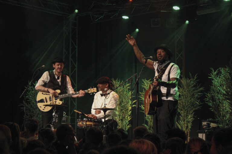 Een band geeft een live optreden met muzikanten op een podium, omringd door groene decoratie en een enthousiast publiek.