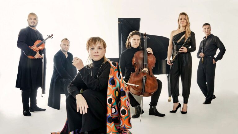 Een groep muzikanten poseert voor een lichtgekleurde achtergrond, met verschillende instrumenten zichtbaar.
