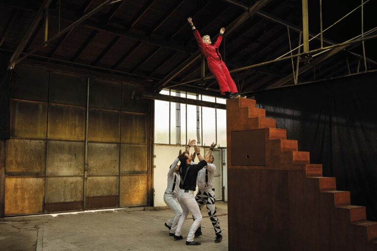 Een vrouw in een rode outfit staat op een trap en steekt haar armen omhoog, terwijl drie andere mensen onderaan de trap staan.