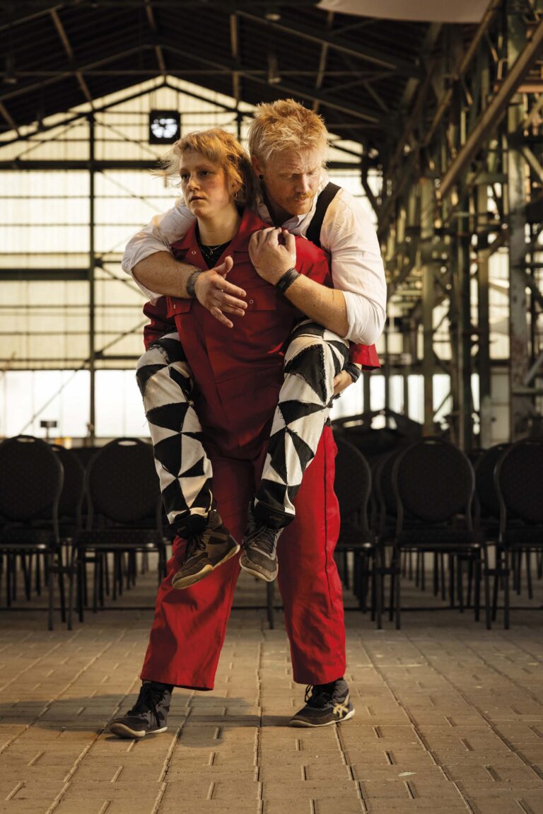 Een man draagt een vrouw op zijn rug in een industrieel ogende omgeving met stoelen op de achtergrond.