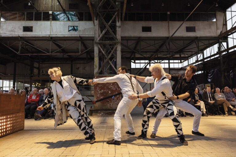 Een groep dansers in opvallende kleding voert een choreografie uit in een grote, industriële ruimte.