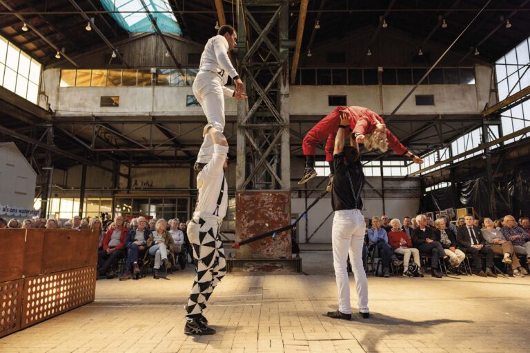 Drie artiesten voeren een acrobatische act uit in een grote zaal, waarbij één performer op een andere balanceert.