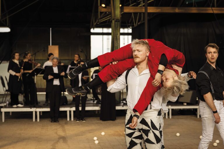 Een performer in een witte blouse en geometrische broek voert een act uit waarbij hij een persoon in een rode outfit over zijn schouder draagt, met een publiek op de achtergrond.