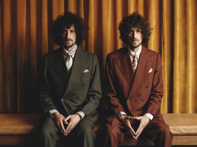 Twee mannen met krullend haar in elegante pakken zitten zij aan zij voor een achtergrond met gordijnen.