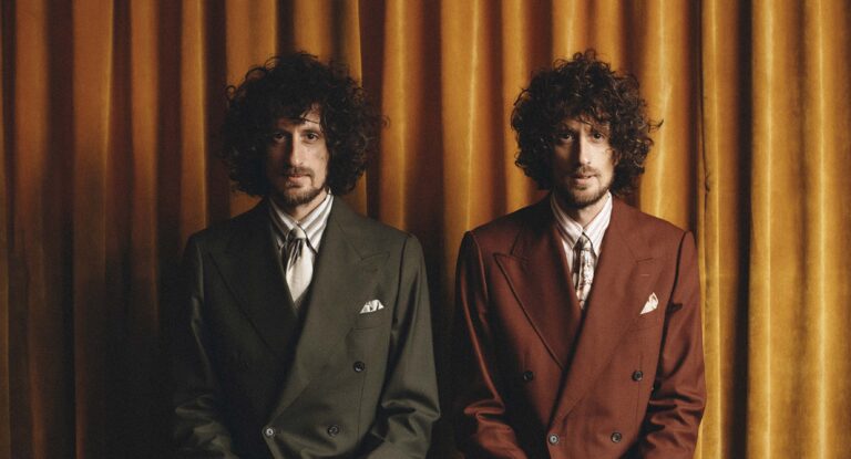 Twee mannen met krullend haar in elegante pakken zitten zij aan zij voor een achtergrond met gordijnen.