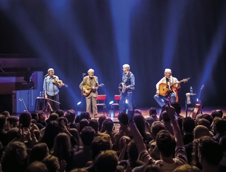 Vier muzikanten optreden op een podium met een enthousiast publiek en kleurrijke lichten.