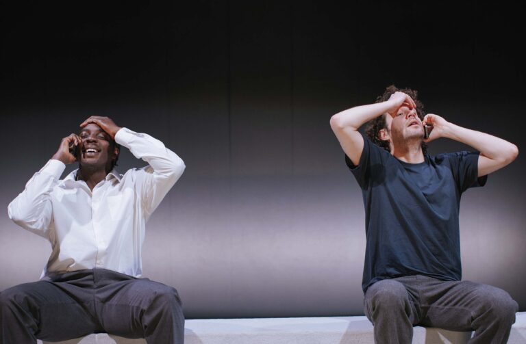 Twee acteurs zitten naast elkaar op een bank en houden hun handen op hun hoofd.