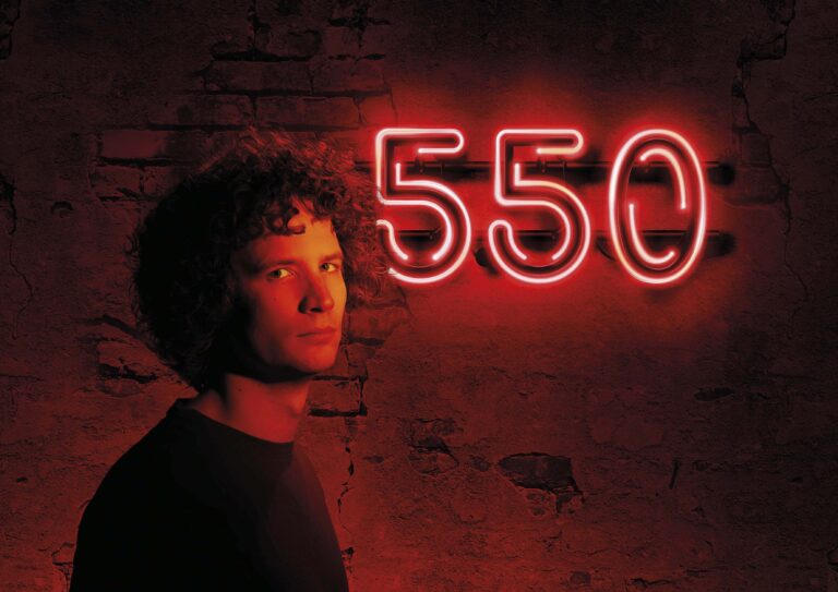 Een persoon met krullend haar staat voor een muur met een neonlicht dat '550' toont.