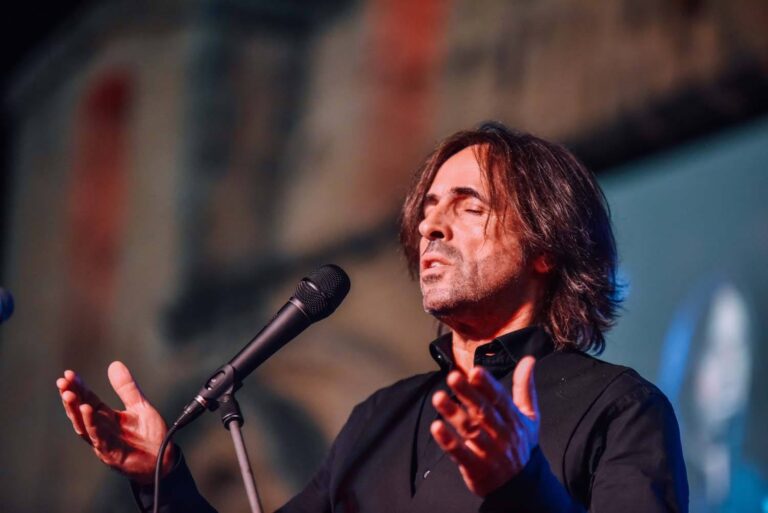 Een man met lang haar staat op het podium en zingt in een microfoon met zijn handen uitgestrekt.